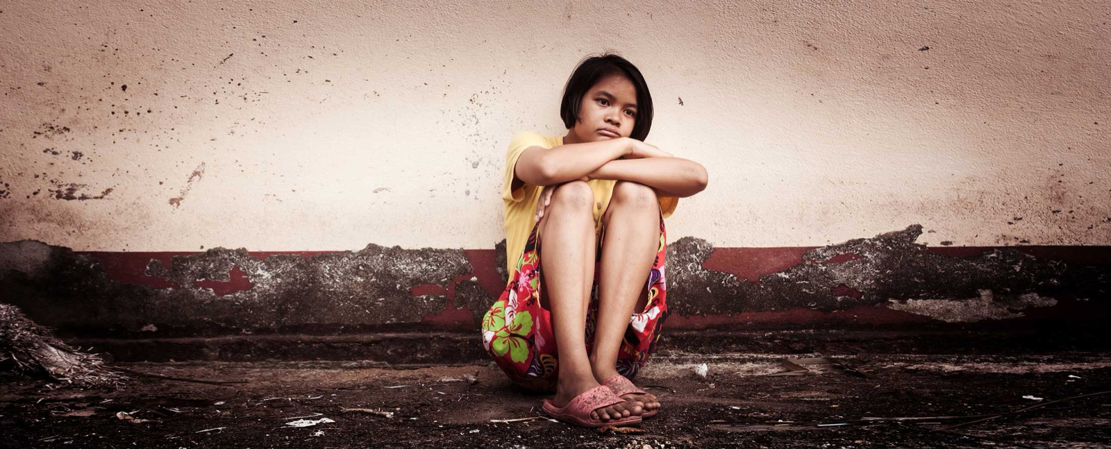 A young girl sits alone against a weathered wall